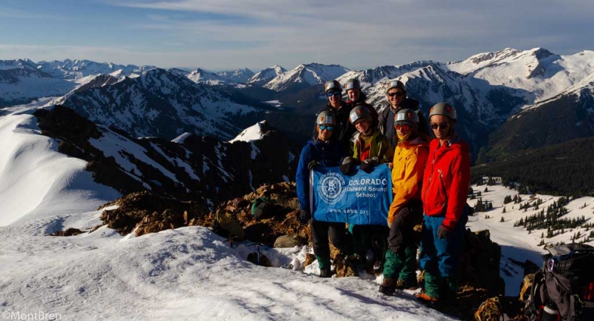 A group of gap year students wearing helmets hold an Outward Bound flag as they stand atop a snowy summit. There are vast snowy mountains in the background.